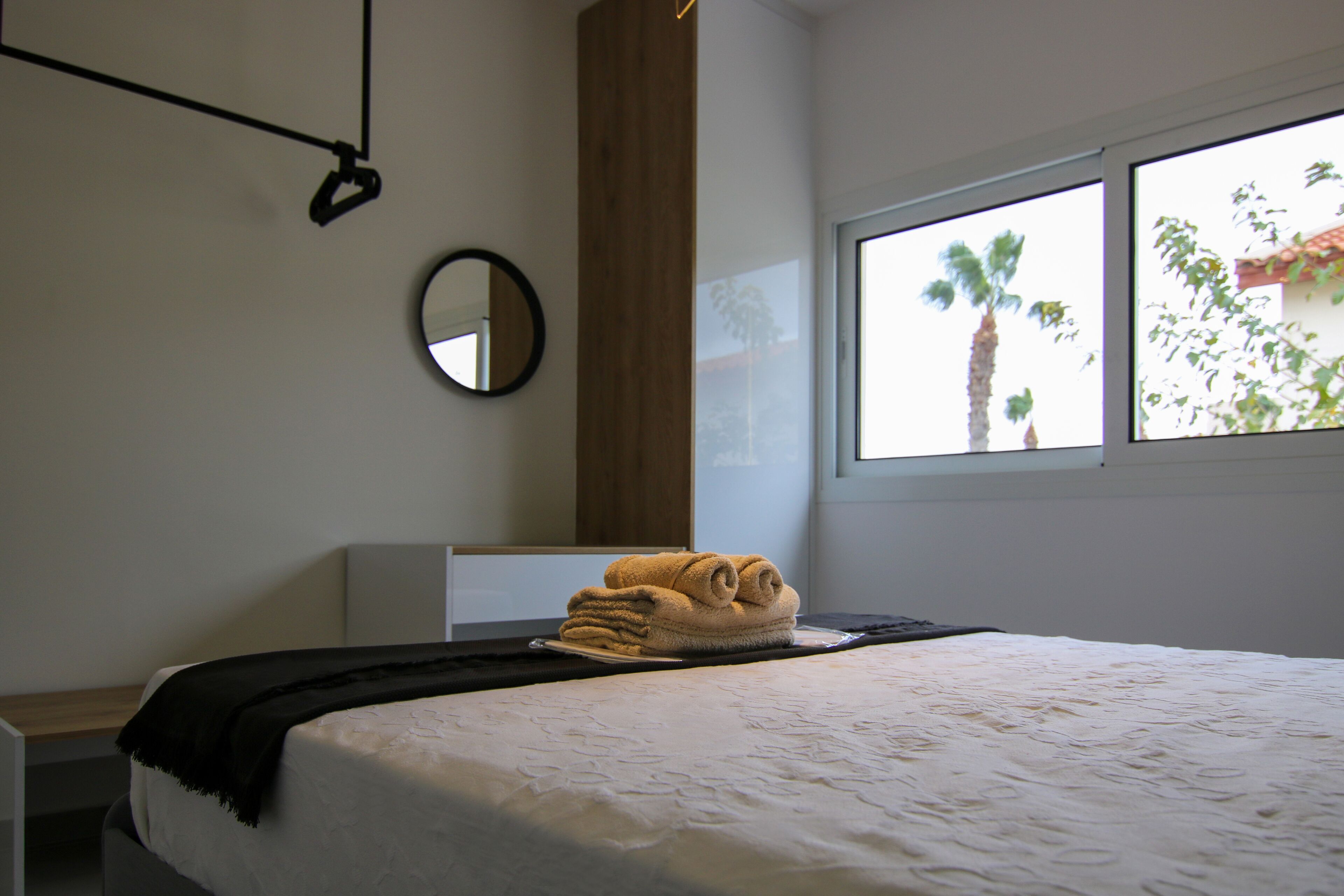 A neatly made bed with a rolled towel, a black throw blanket, and a wooden headboard. A round mirror hangs on the wall, and a window with a view of palm trees is visible.