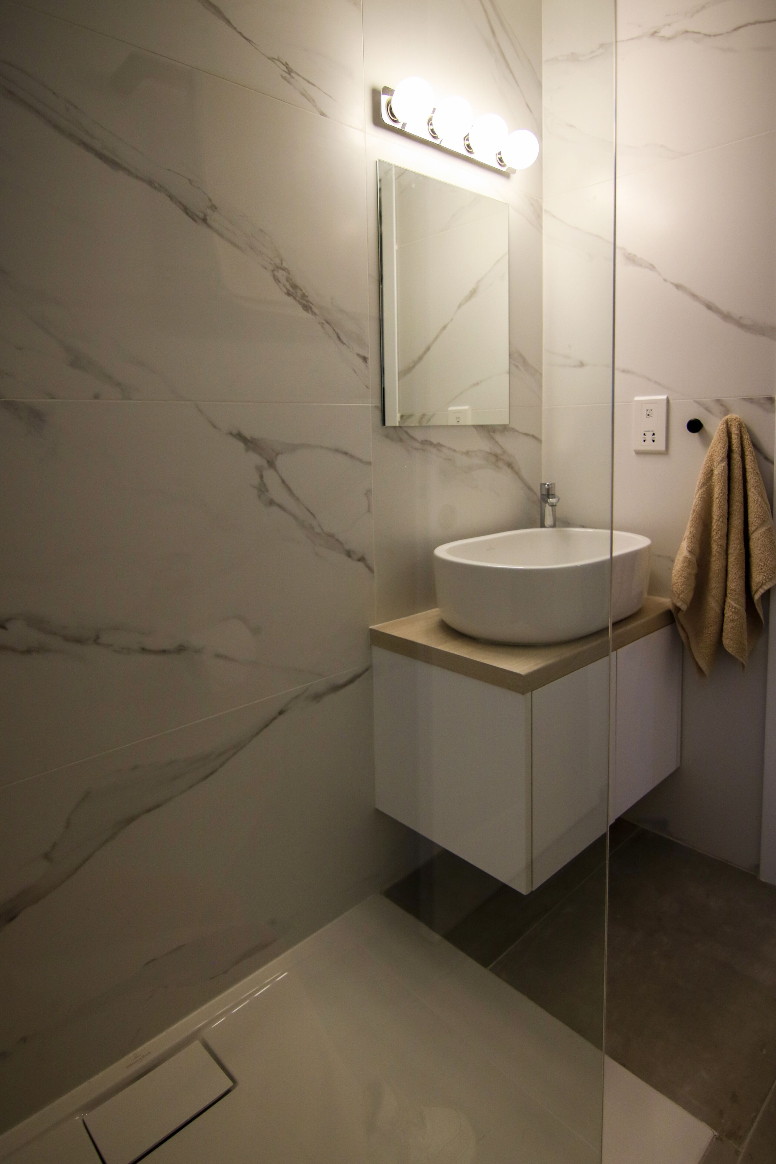 Une salle de bain avec un mur en marbre, un lavabo blanc, un miroir avec éclairage intégré et une serviette accrochée au mur.