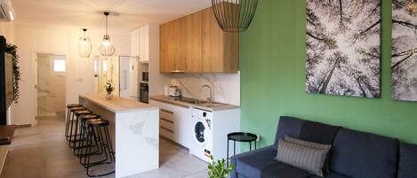 Een moderne keuken met een centraal eiland, barstoelen en een wasmachine. Er is een bank en een salontafel met een plant.