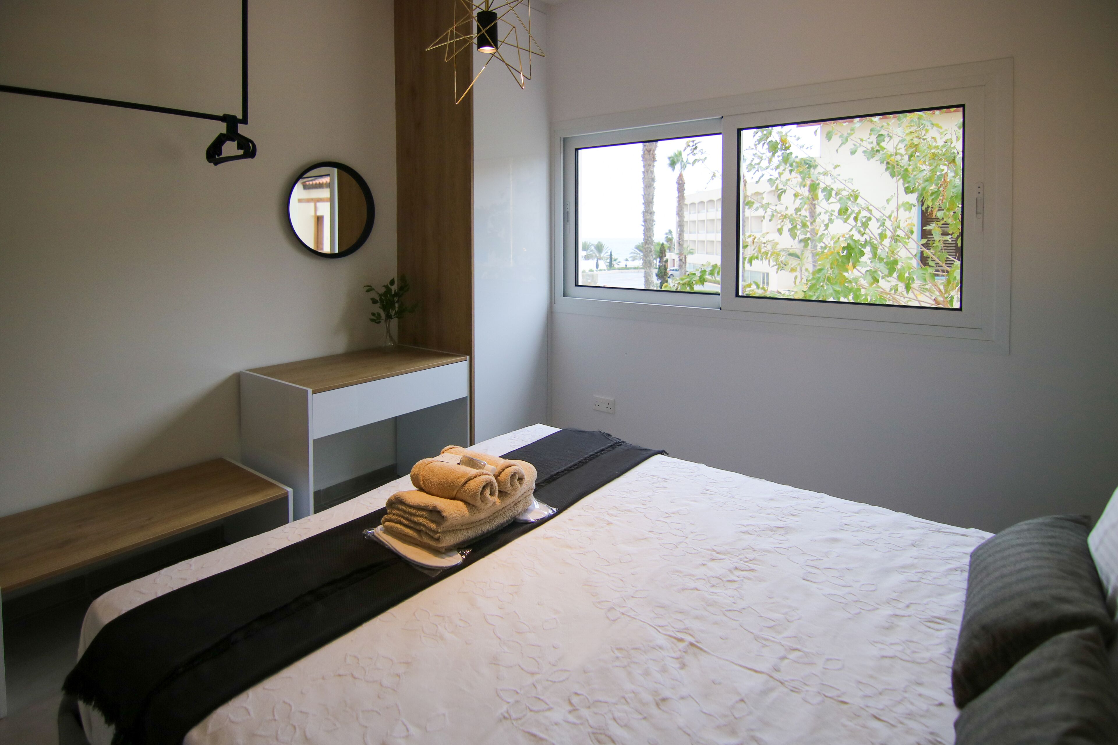 Une chambre moderne avec un lit bien fait, une table de chevet, un miroir et une fenêtre donnant sur des arbres et un immeuble.