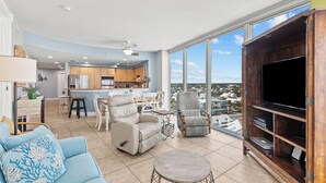 A spacious living room with a large TV, comfortable seating, a kitchen in the background, and a view of the city through large windows. - Island Tower 1101 by Liquid Life (Gulf Shores)