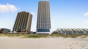 A tall, modern hotel with a curved facade and a flat roof, situated on a sandy beach with other buildings in the background. - Island Tower 1101 by Liquid Life (Gulf Shores)