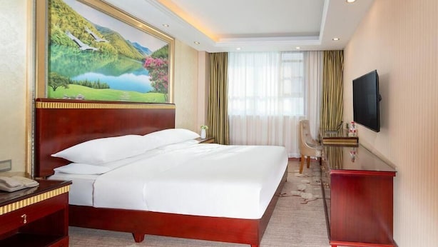 A hotel room with a large bed, a desk with a telephone, a TV mounted on the wall, and a painting of a landscape. - Vienna Hotel (Huizhou Zhongkai Chenjiang Avenue) (Huizhou)
