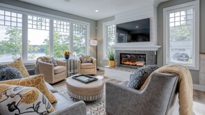 A spacious living room with a fireplace, comfortable seating, large windows, and a flat-screen TV. - Traverse City Lakemore Resort,  New Luxury Lakefront Home on Arbutus Lake (Traverse City)