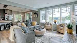 A spacious living room with a grey sofa, ottoman, and two armchairs. A kitchen area is visible with a refrigerator and bar stools. Large windows provide natural light and a view of greenery outside. - Traverse City Lakemore Resort,  New Luxury Lakefront Home on Arbutus Lake (Traverse City)