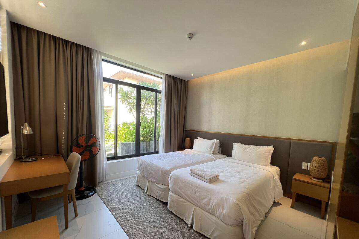 A hotel room with two beds, a desk, a chair, a fan, and a window with curtains.