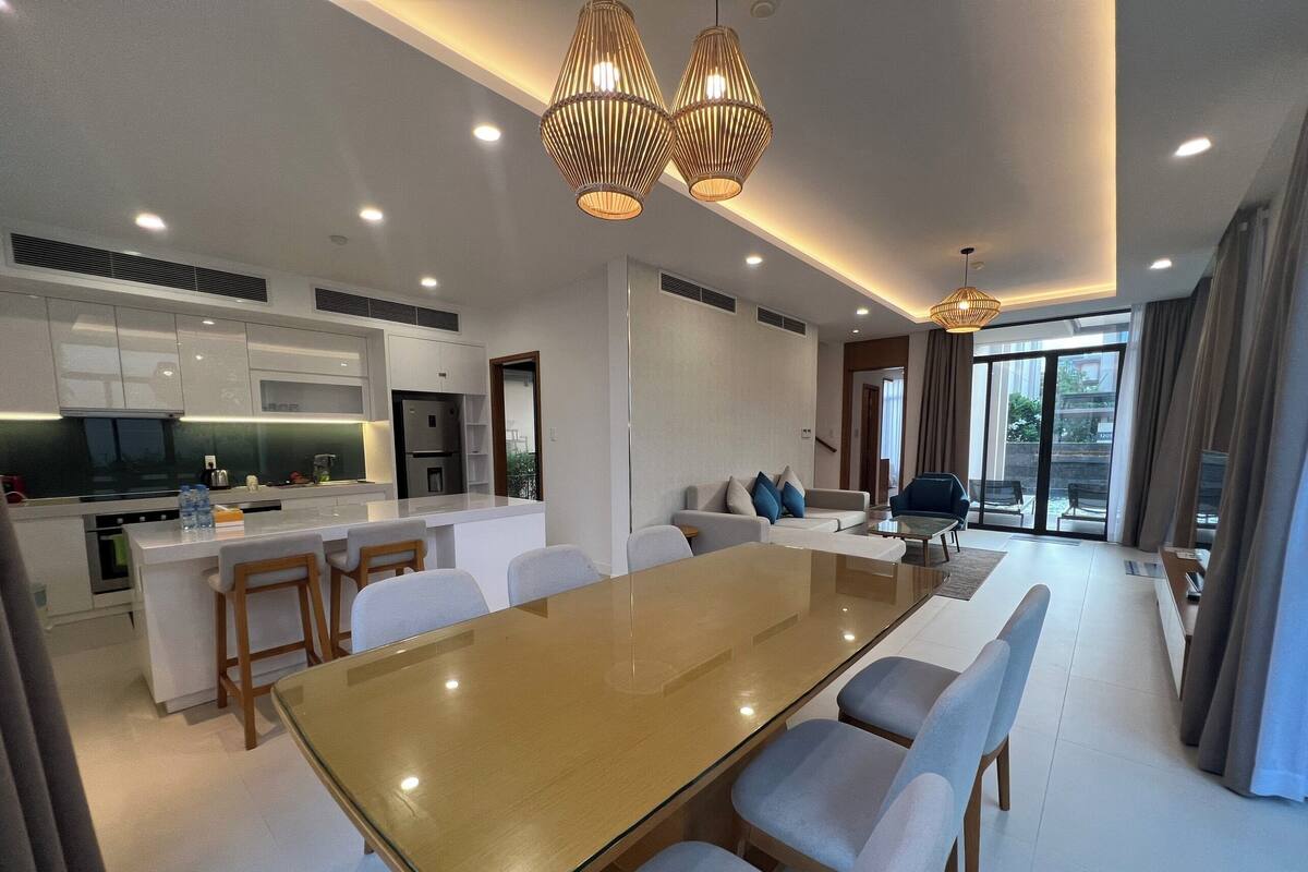 A modern kitchen with white cabinets, a central island, and stainless steel appliances. There is a dining area with a glass-top table and chairs.