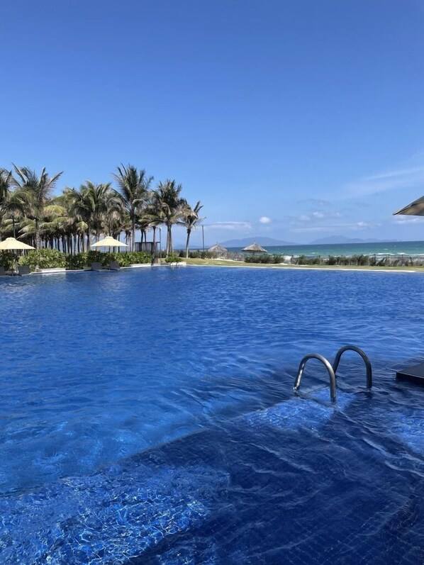 A clear blue pool with lounge chairs and umbrellas, surrounded by palm trees and a view of the ocean. - Suite & Pavilion Thien Kim (Cam Lam)