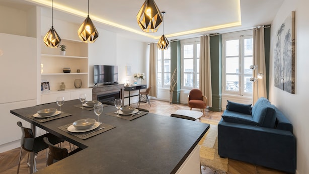 A modern living room with a sofa, a dining area with a table set for two, and a kitchen area with a TV and shelves. - Le Confident - Grand T2 au coeur de Lyon (Lyon)