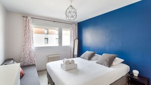 A bedroom with a large bed, a radiator, a window with curtains, a bedside table, and a blue accent wall. - LORGERIL - appartement 2 chambres proche du Mail (Rennes)