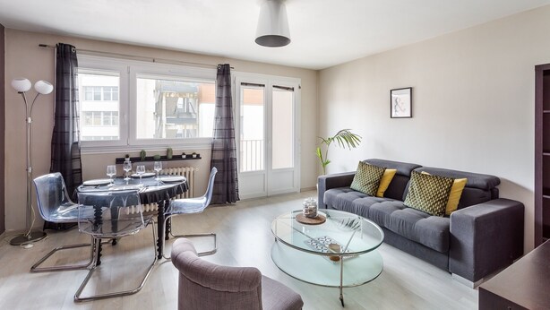 A modern living room with a dining area, a glass coffee table, a grey sofa with patterned cushions, and a large window with curtains. - LORGERIL - appartement 2 chambres proche du Mail (Rennes)