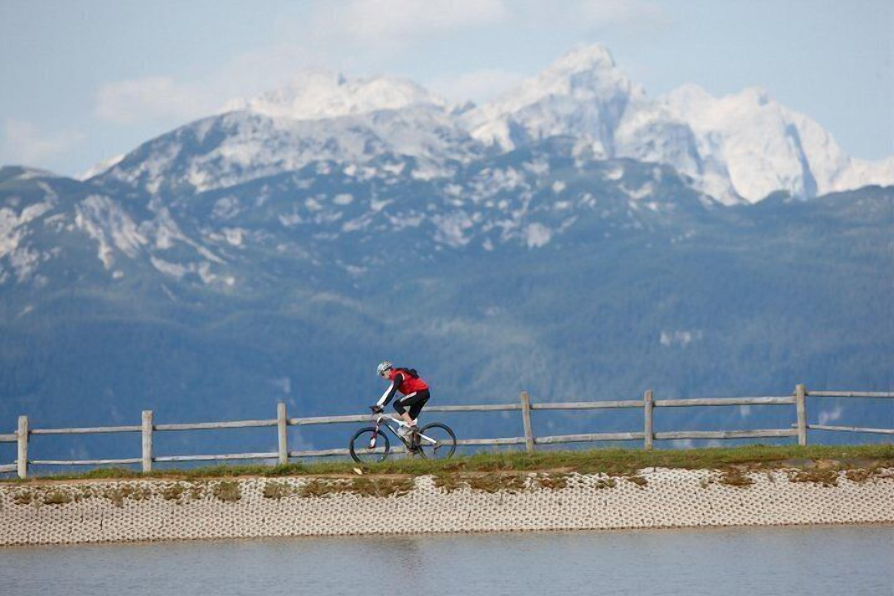 A person riding a bicycle on a paved path with a wooden fence and a body of water in the foreground, and snow-capped mountains in the background.
