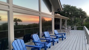 Een houten terras met blauwe Adirondack-stoelen, grote ramen met uitzicht op een zonsondergang, en een overdekt gedeelte met stoelen en een barbecue.