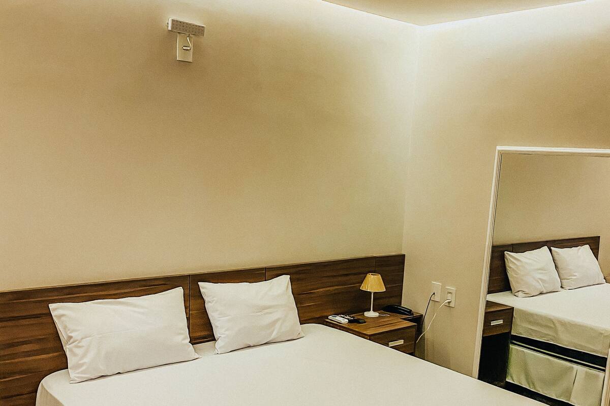 Un lit bien fait, avec du linge de lit blanc et deux serviettes enroulées, une tête de lit en bois, une table de chevet avec une lampe et un miroir au mur.