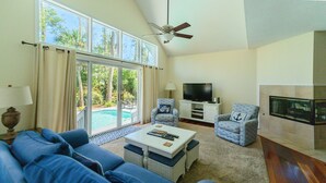 A living room with a large window, a ceiling fan, a flat-screen TV, and a fireplace. - 3rd row updated home in Palmetto Dunes Resort, large private pool & hot tub (Hilton Head Island)