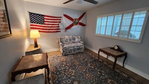 A room with a patterned rug, a floral armchair, a small table with a lamp, and two flags on the wall. - Great location with ocean view  . (Venice)