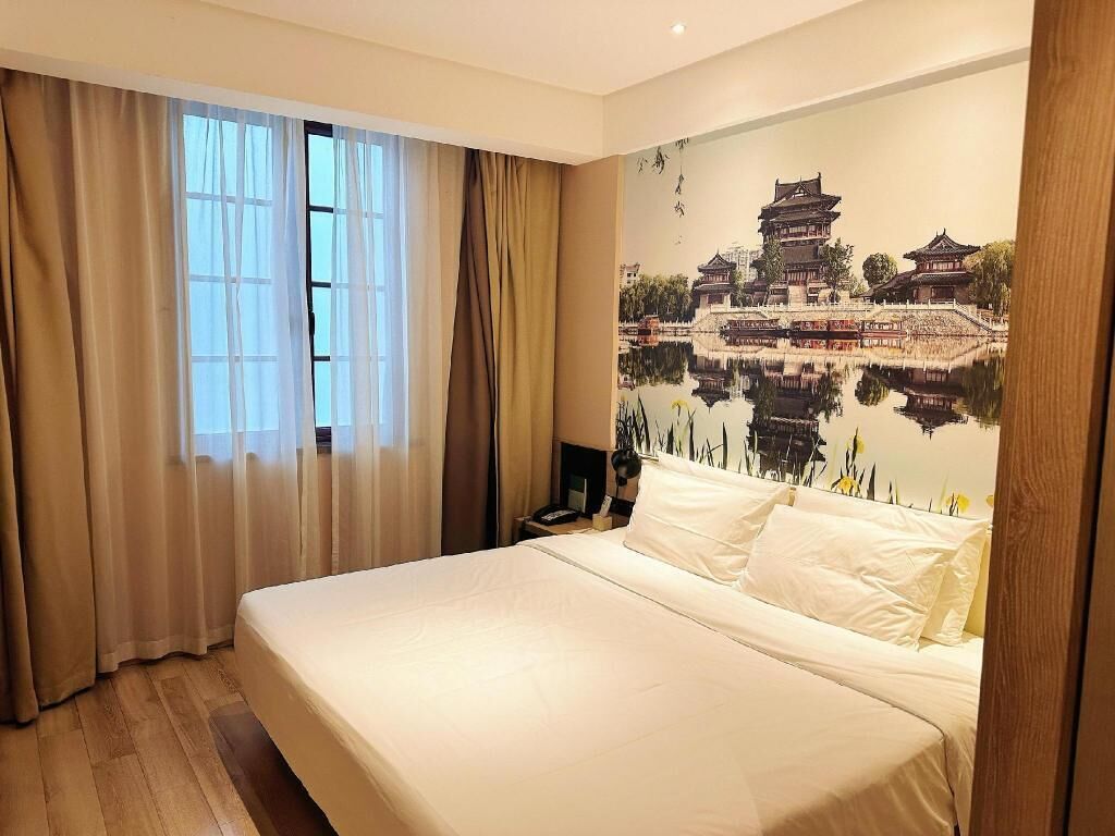 A hotel room with a large bed, beige curtains, a nightstand, and a wall mural of a traditional building.