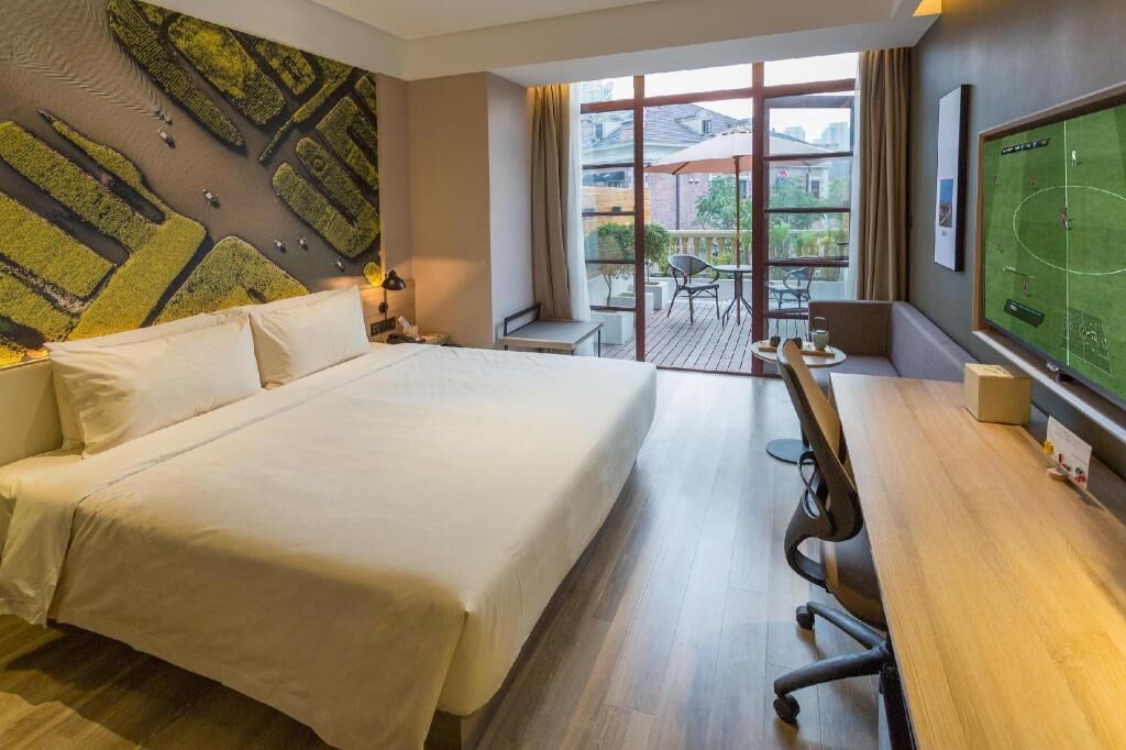 A hotel room with a large bed, a desk with a chair, a small table with a chair, a large window with a view of buildings, and a wall-mounted TV.