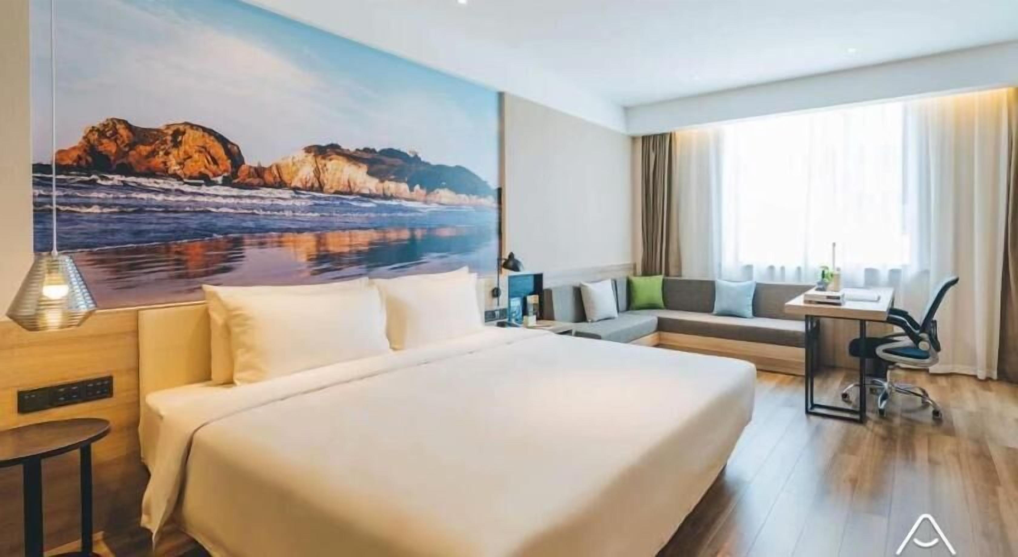 A modern hotel room with a large bed, a seating area with a sofa and armchairs, a desk, and a large window with curtains.