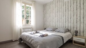 A bedroom with a bed, a nightstand, a window with curtains, and a wall with vertical lines. - Villa 6km from St Rémy de Provence with swimming pool for 2 to 10 people (Eyragues)