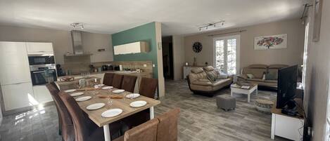 A spacious living area with a dining table set for four, a sofa, a TV, and a kitchen in the background.