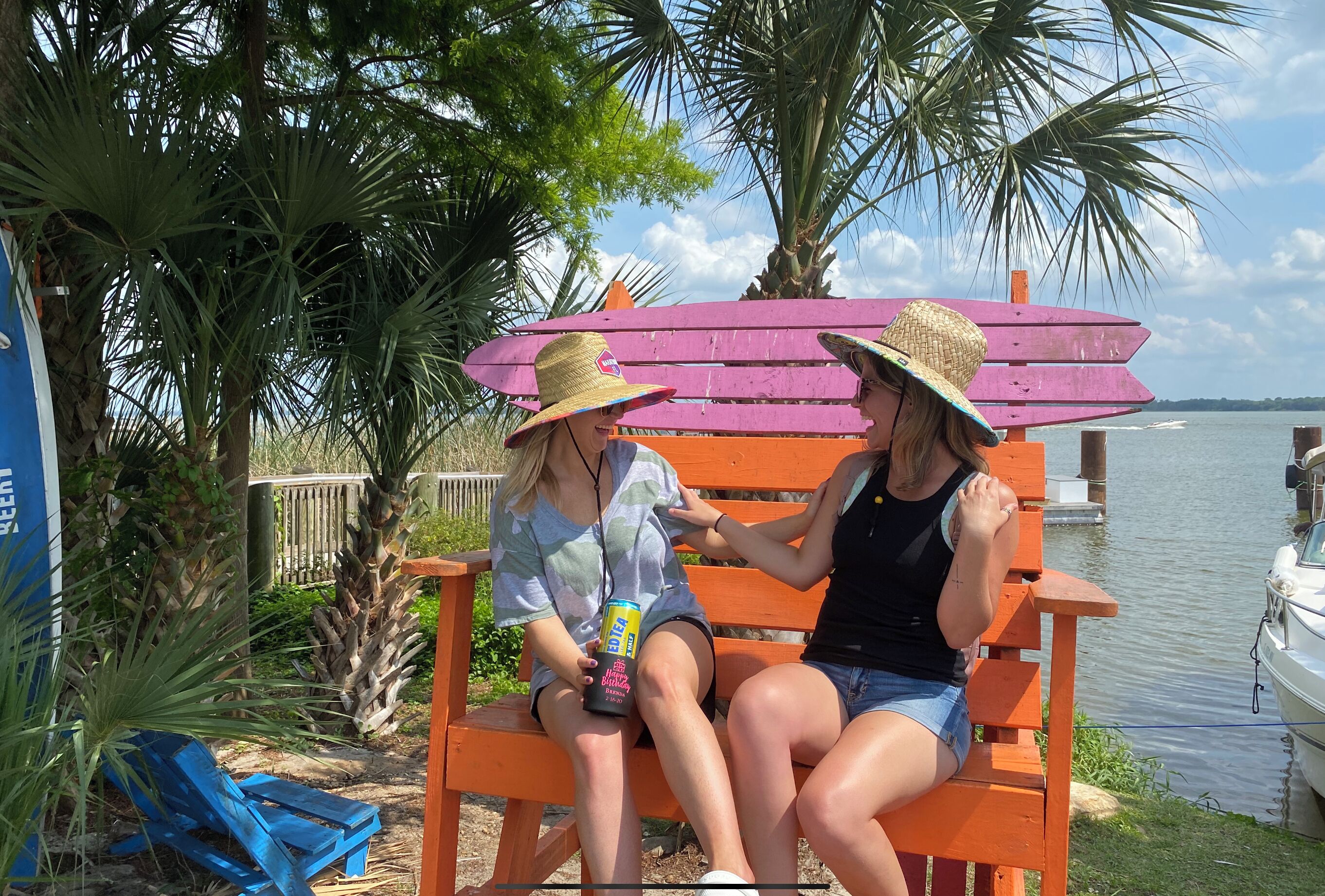 Two people sitting on a bench by a lake, with a boat and palm trees in the background.