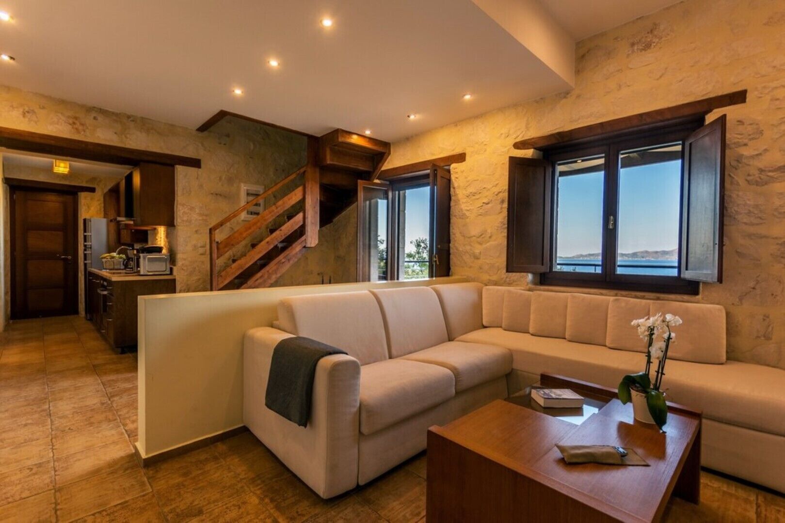 Una sala de estar moderna con un sofá seccional color beige, una mesa de centro de madera y vista al mar a través de ventanas grandes.