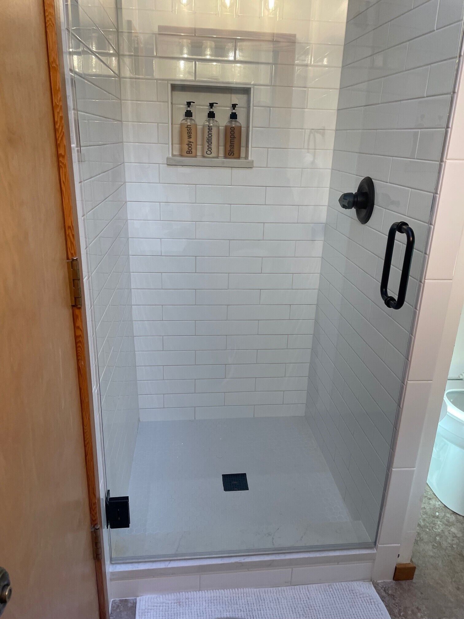 A glass-enclosed shower with white tiled walls and a shelf with toiletries.