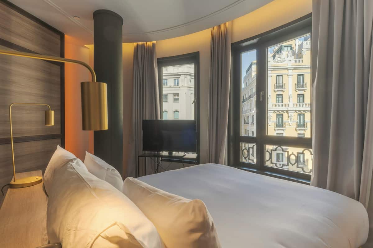 A hotel room with a bed, a TV, a lamp, and a large window with a view of buildings.