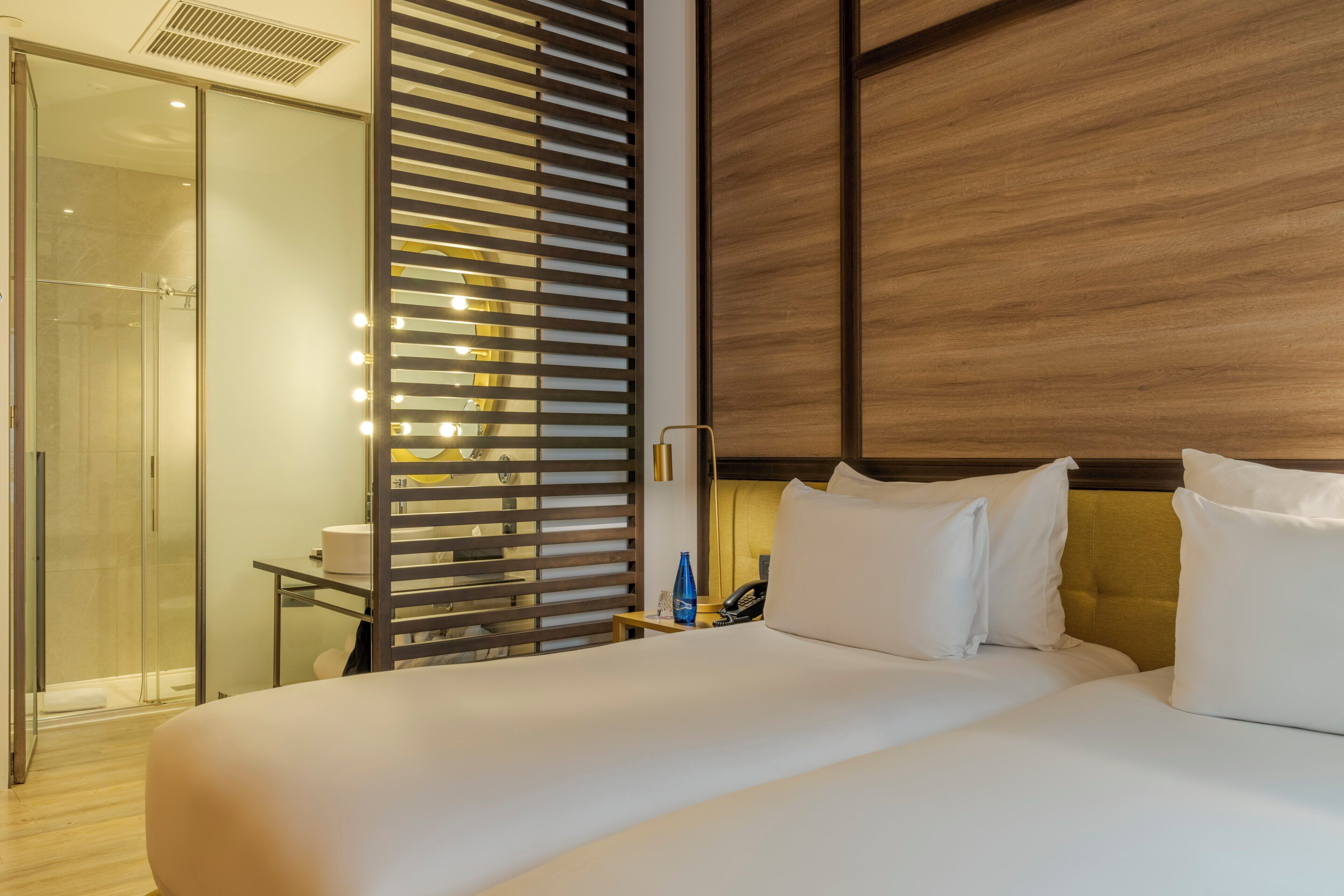 Habitación de hotel con una cama grande, cabina de ducha de vidrio, cabecero de madera y mesita de noche con un teléfono y una botella.