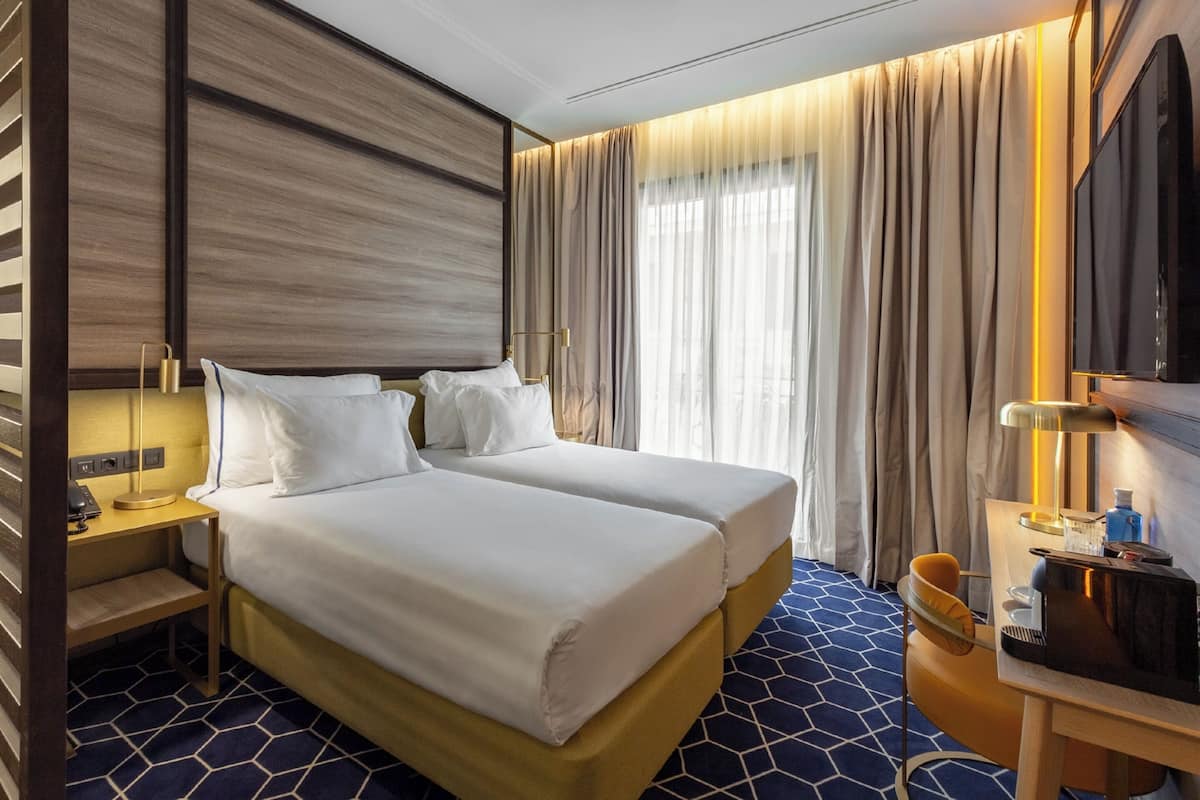 A hotel room with two beds, a desk, a chair, a TV, and a large window with curtains.