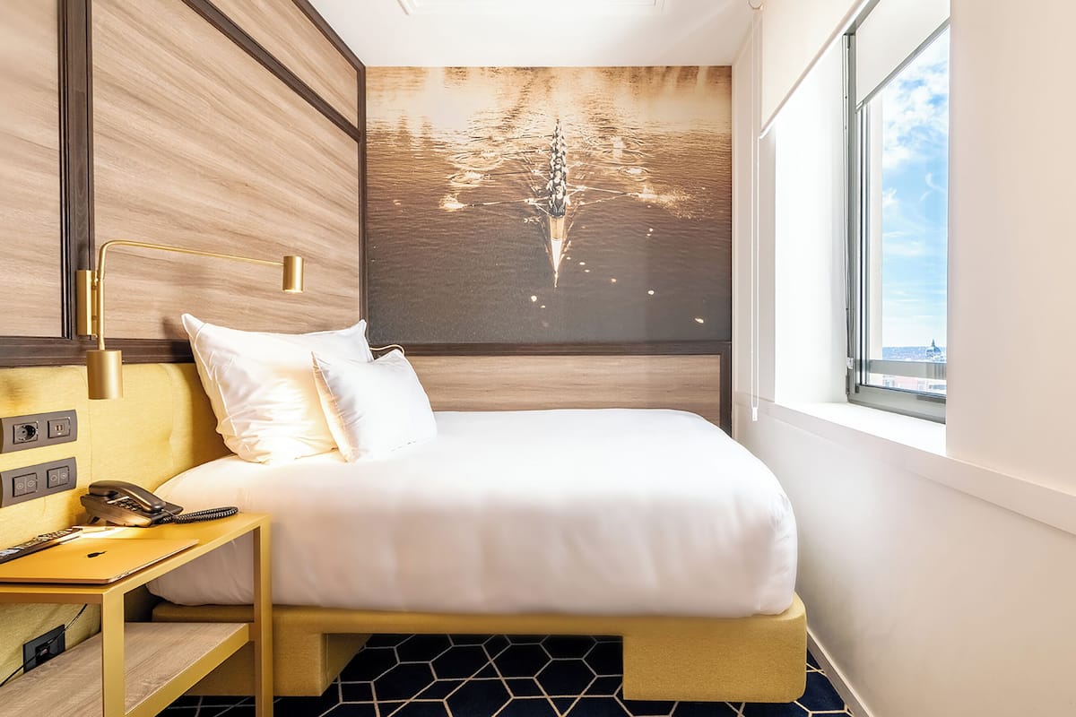 A modern hotel room with a bed, bedside table, lamp, and a cityscape view through the window.