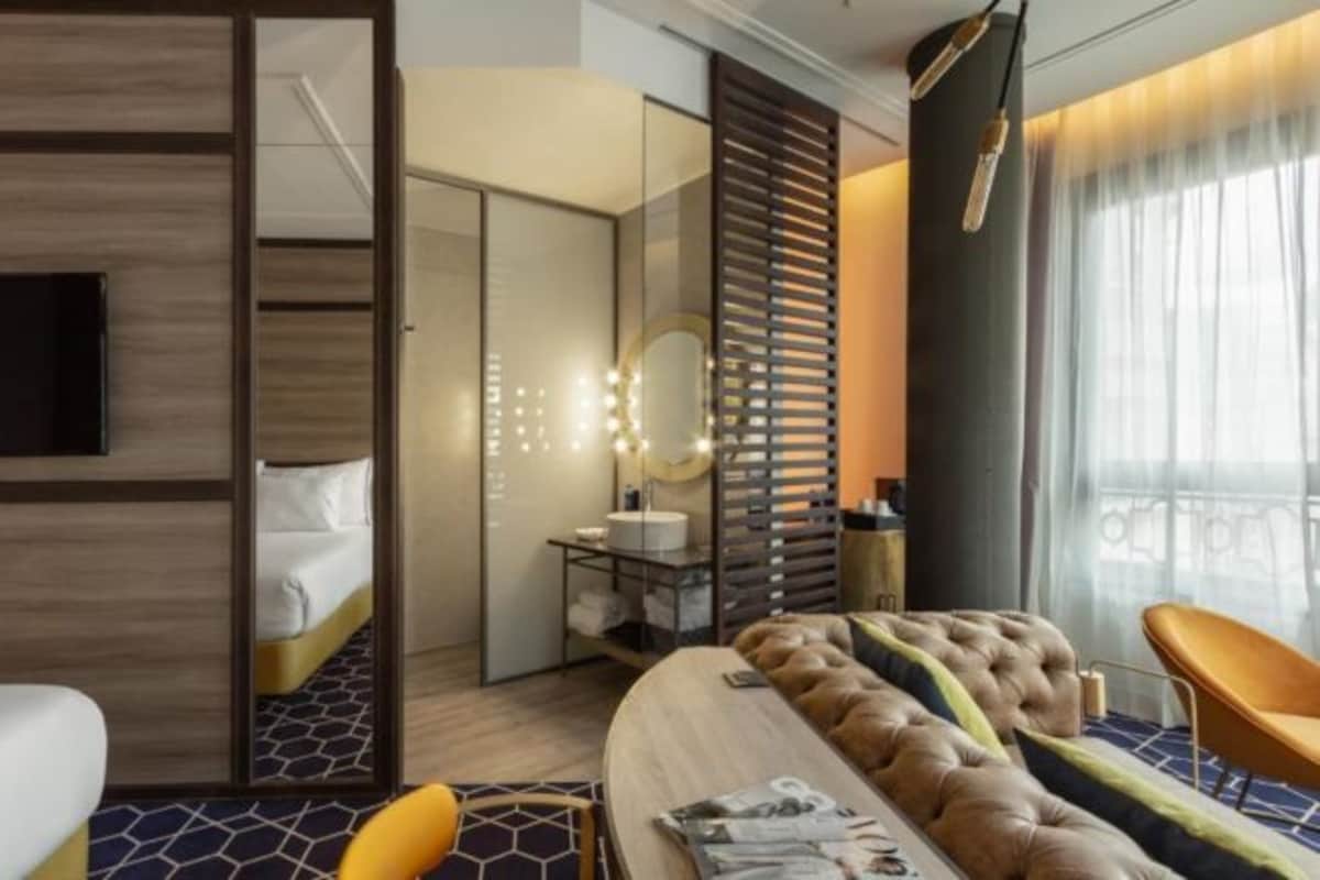 Habitación de hotel con cama, zona de estar con sofá y sillón, tocador con espejo y ventana con cortinas.