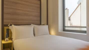 Habitación de hotel con cama, mesita de noche, una botella y ventana con vista a edificios.