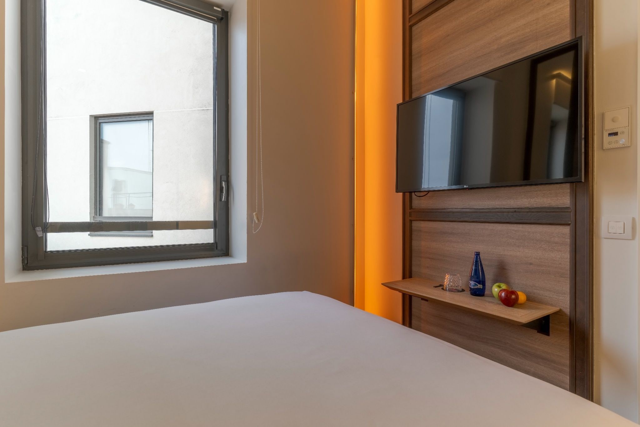 Una camera d'albergo con un letto, una finestra, una TV montata su un pannello di legno e un piccolo ripiano con una bottiglia e della frutta.