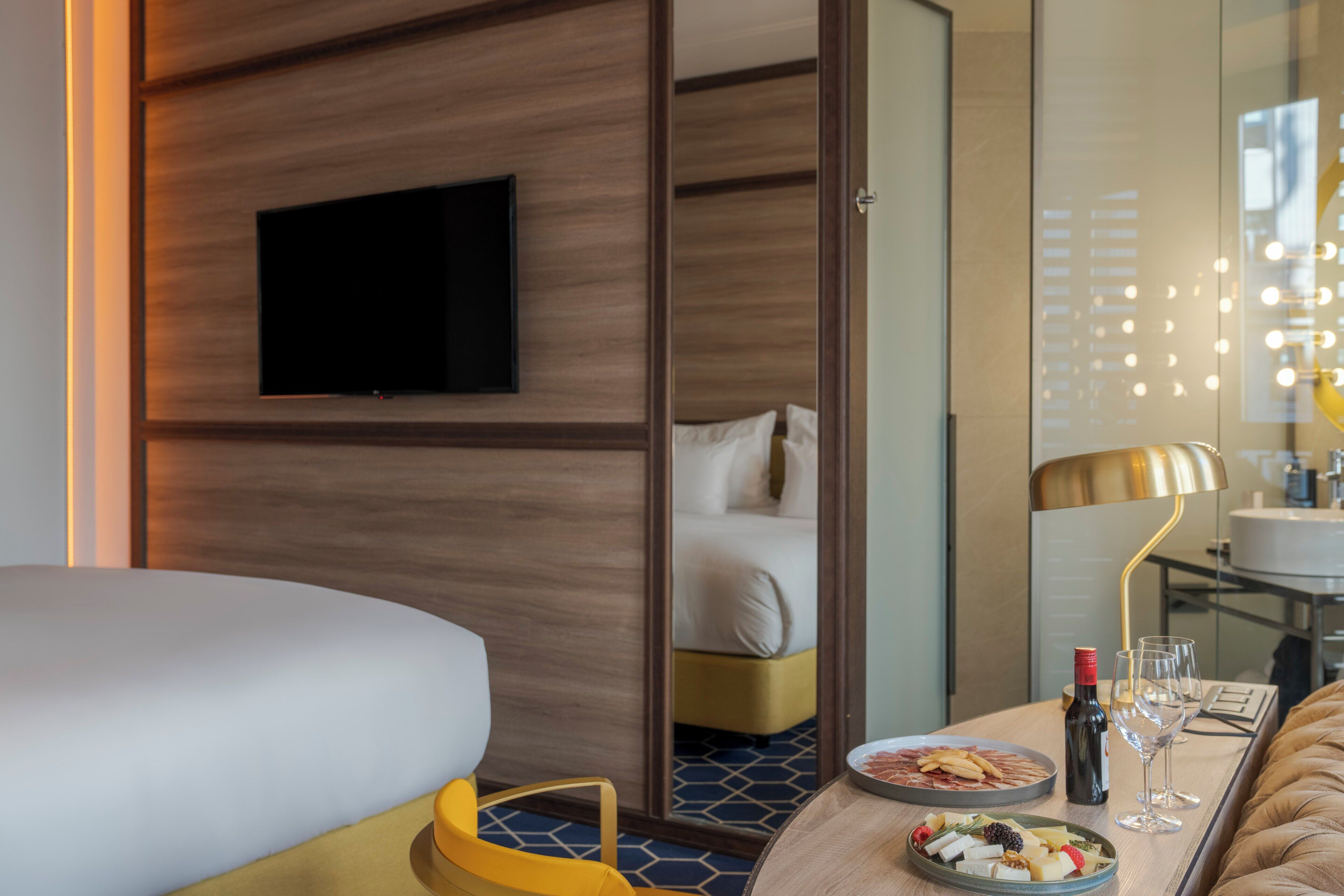 Una camera d'albergo con un letto, una TV a parete, un tavolo da pranzo con un piatto di cibo, una bottiglia di vino e una lampada.