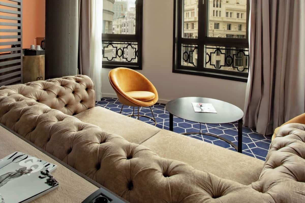 A modern hotel room with a large tufted sofa, a round table, and a unique ceiling light fixture.