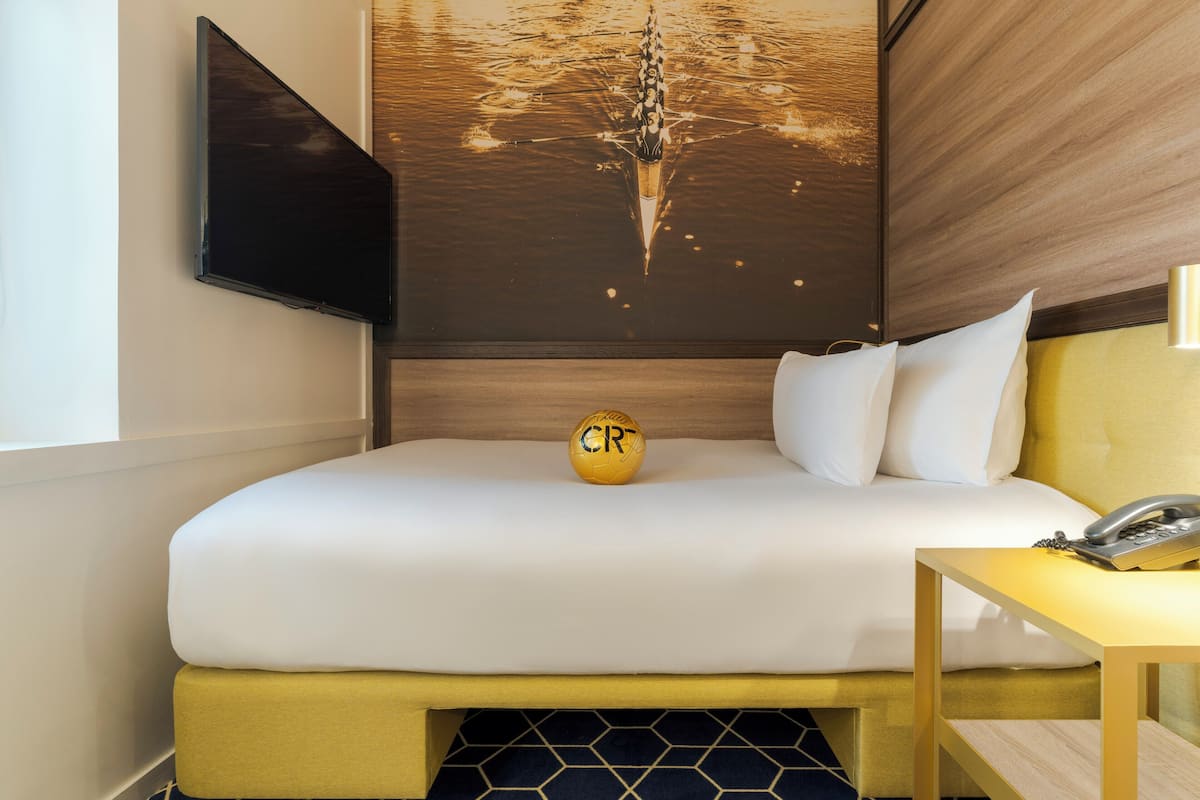 A hotel room with a bed, a bedside table with a telephone, a wall-mounted TV, and a decorative wall piece.