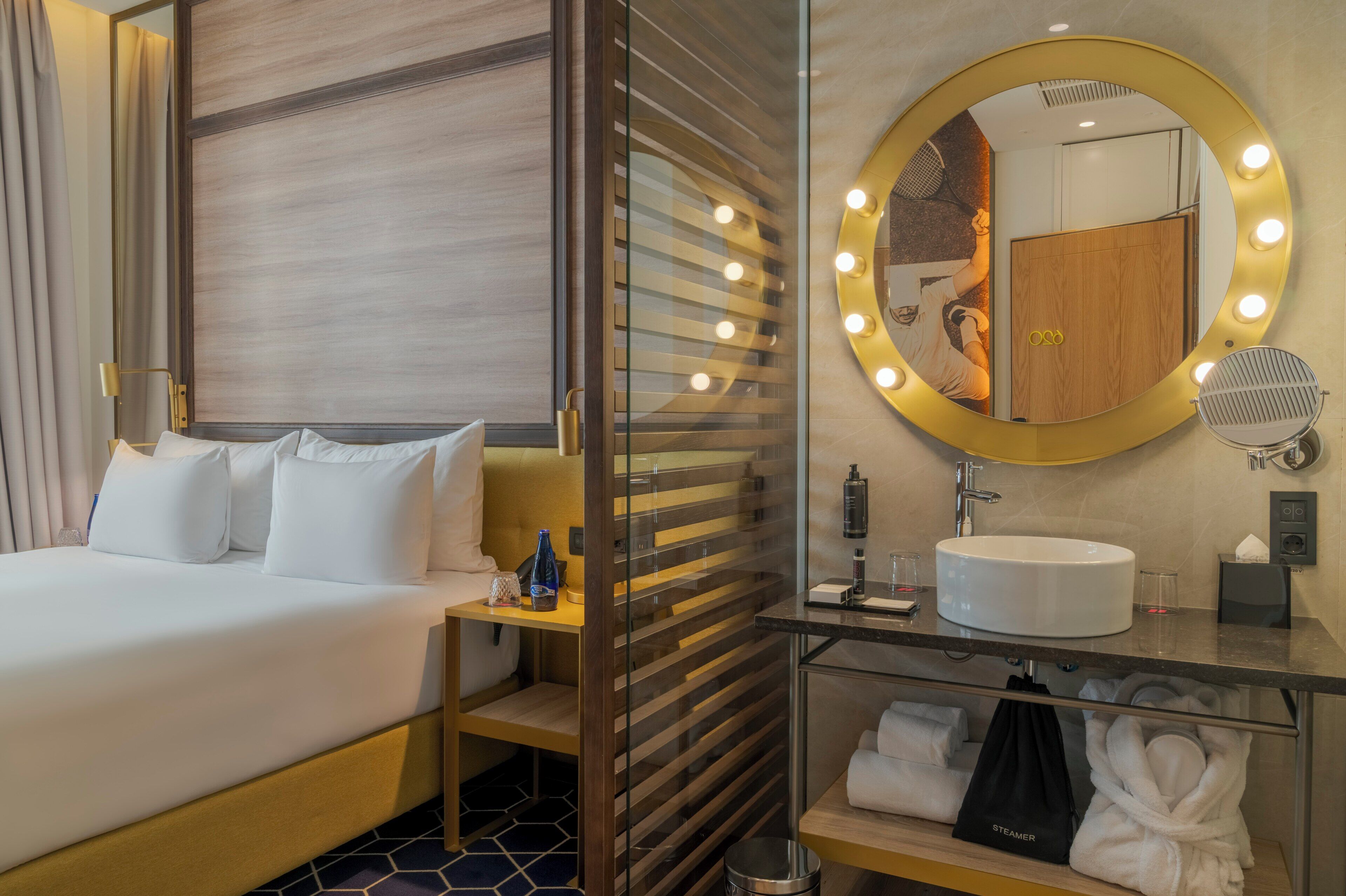 A hotel room with a bed, a vanity with a mirror and lights, a sink, and a robe rack.