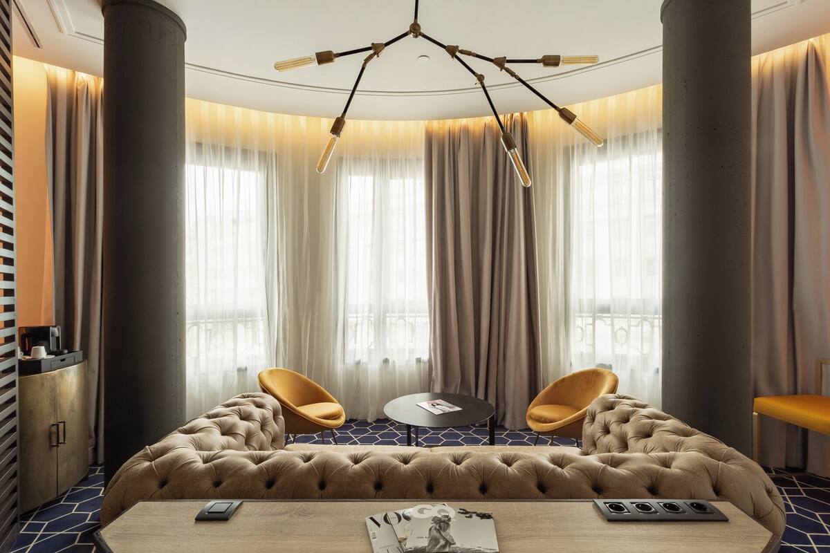 A modern hotel room with a tufted sofa, two yellow chairs, a round table, and a unique ceiling light fixture.