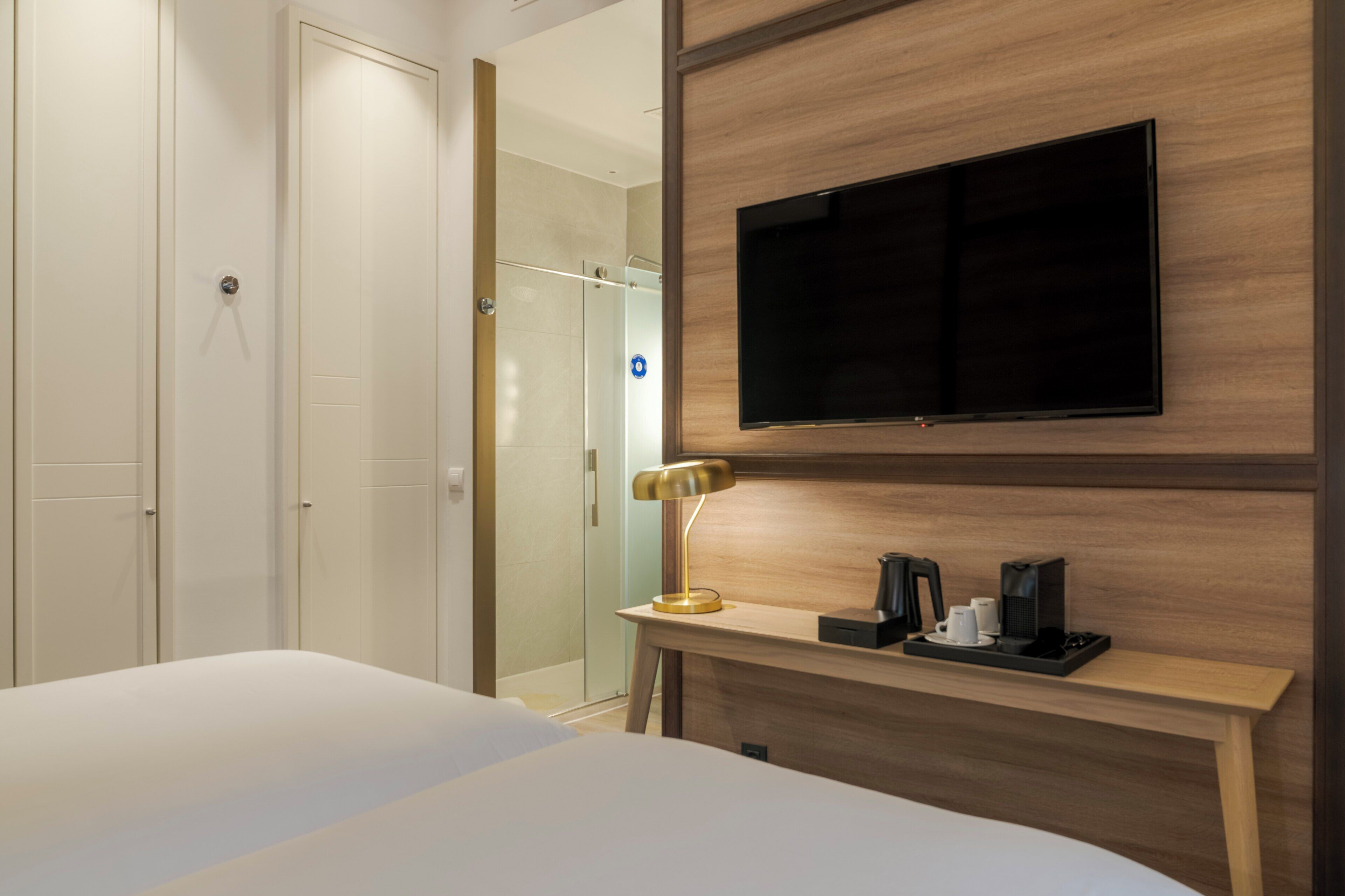 A hotel room with a bed, a TV mounted on the wall, a bedside table with a lamp, and a shower area.