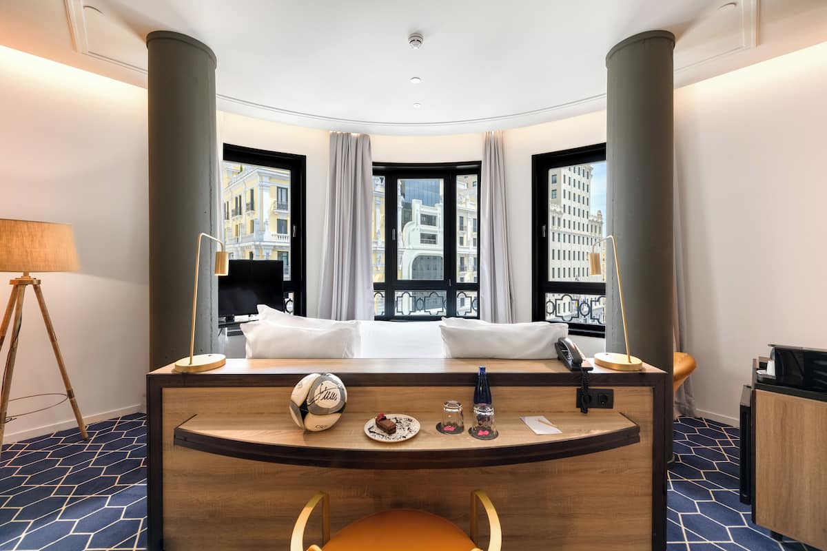 A hotel room with a curved desk, a bed, a lamp, a phone, and a view of buildings through large windows.