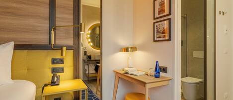 A hotel room with a bed, bedside table with a lamp, a small desk with a chair, a mirror with lights, and a shower area.