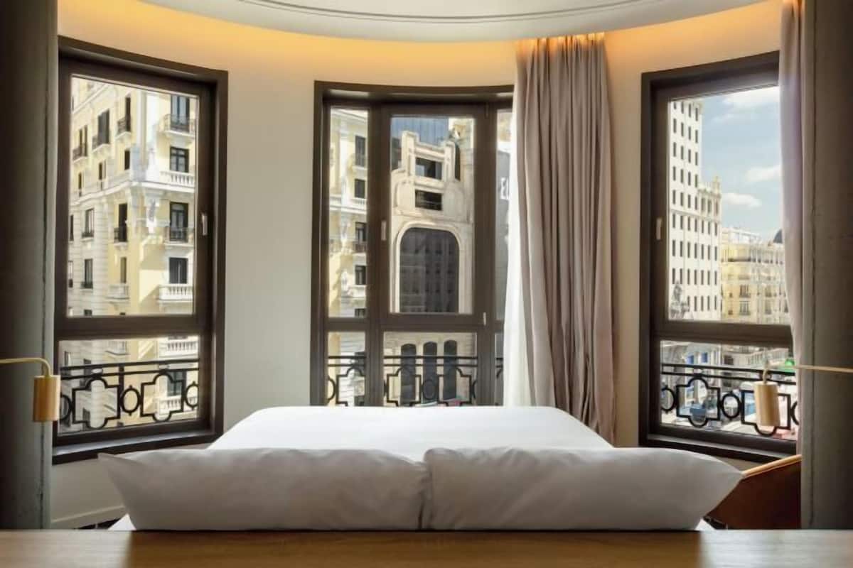 A hotel room with a bed, large windows, and a view of city buildings.