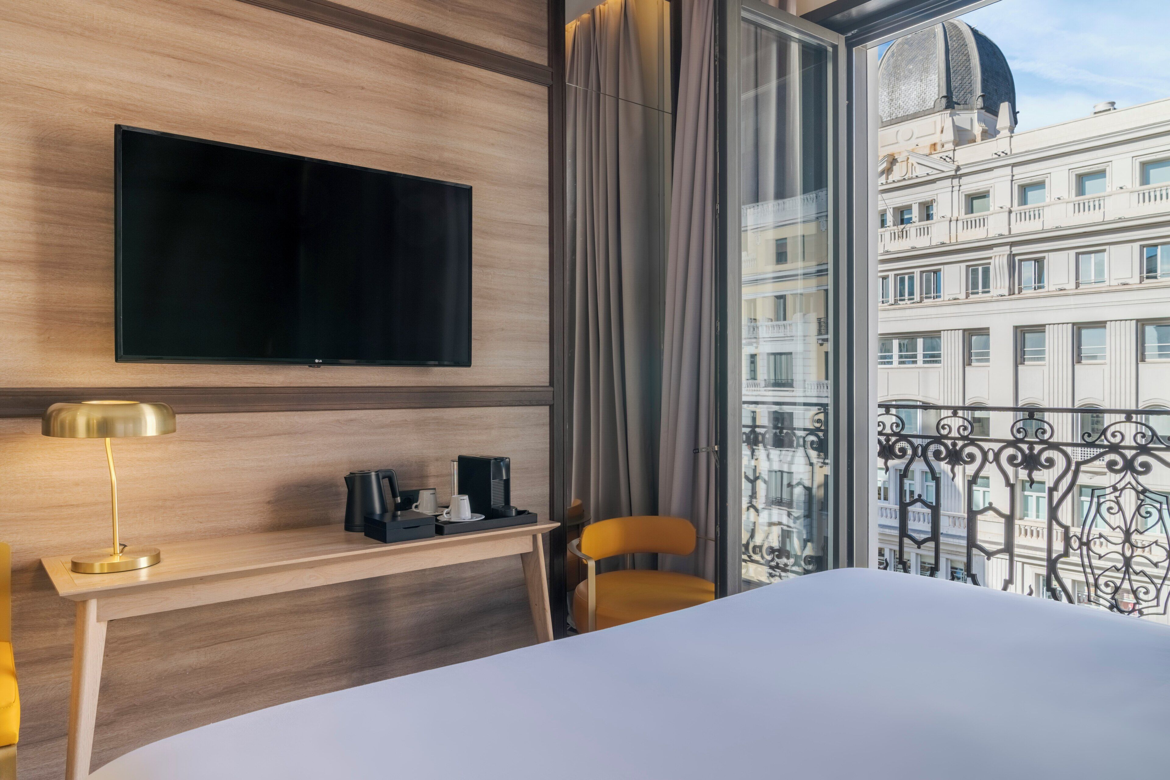 Ein Hotelzimmer mit Bett, Schreibtisch, Kaffeemaschine, Fernseher und Blick auf ein Gebäude durch ein Fenster.