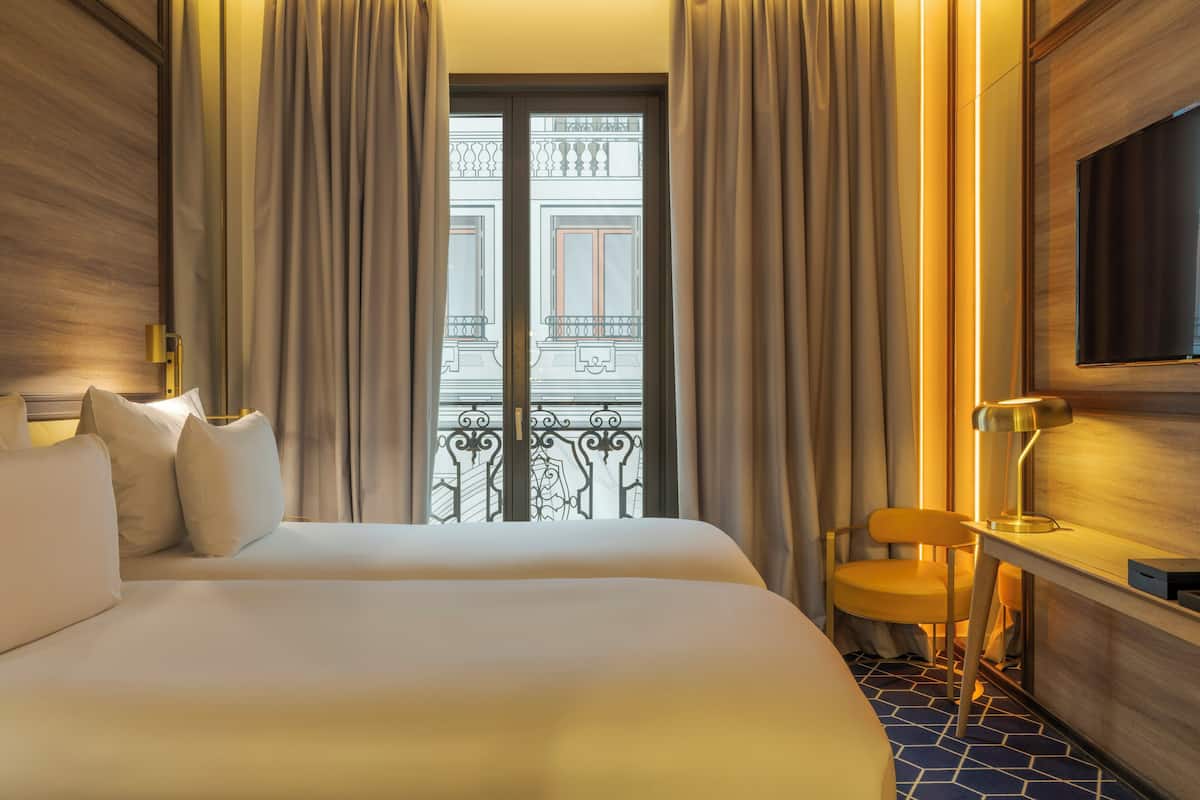 A hotel room with a bed, a chair, a desk with a lamp, a TV, and a view of a balcony with curtains.