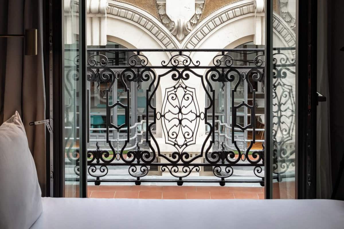 A hotel room with a balcony featuring ornate wrought iron railings and a view of a classical building with columns and arches.