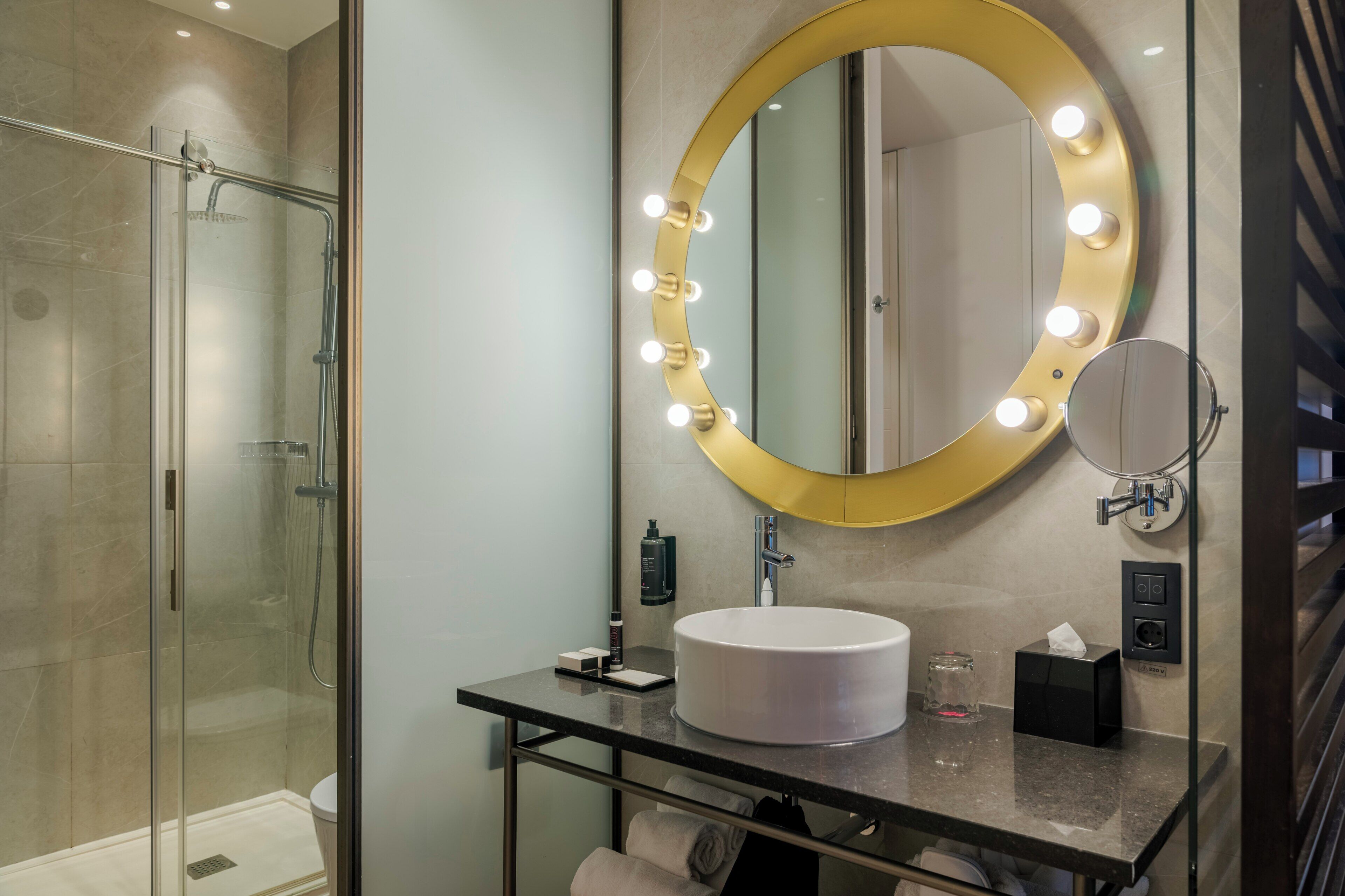 A modern bathroom with a glass-enclosed shower, a vanity with a round mirror and lights, and a sink with toiletries.