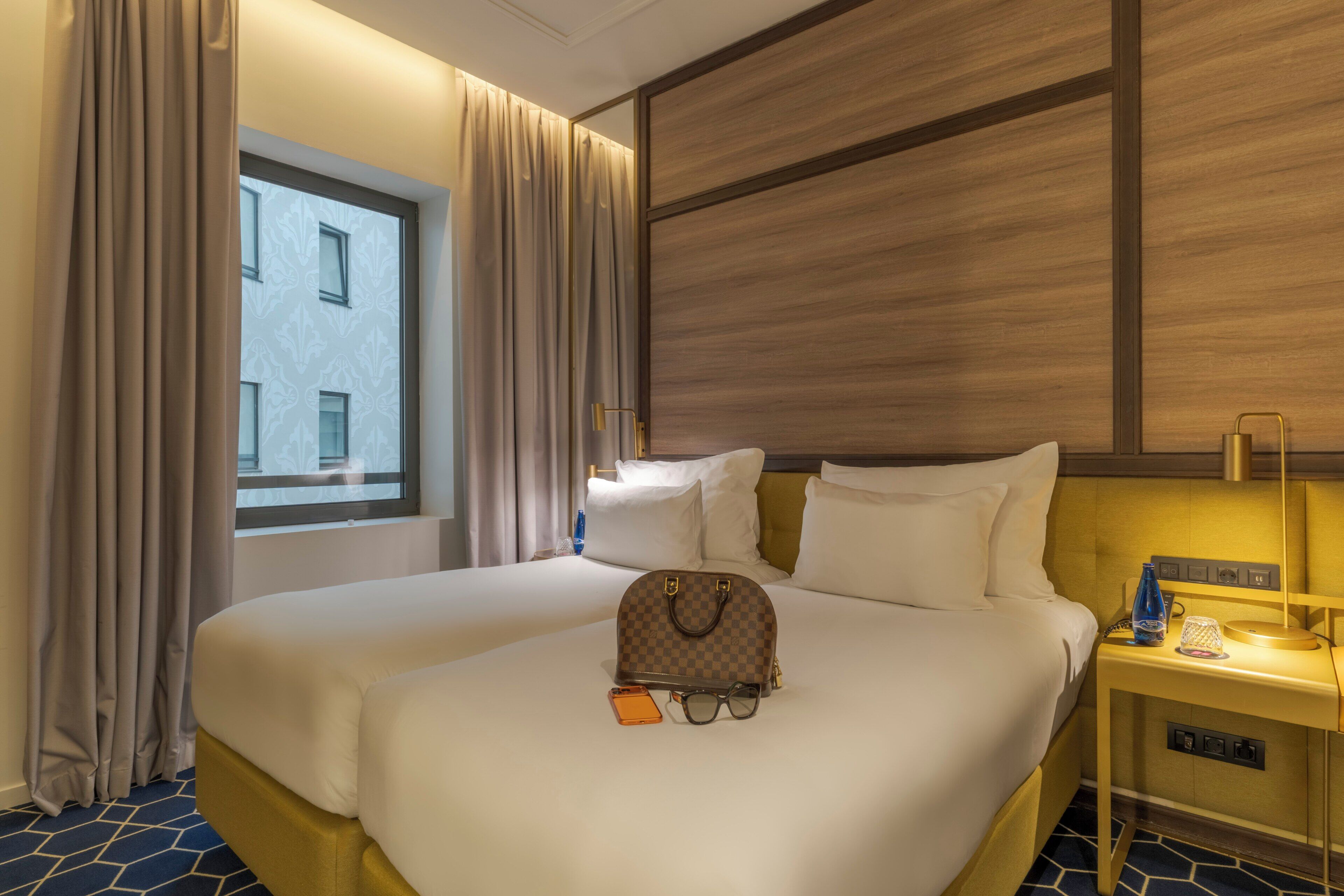 Habitación de hotel con una cama grande, una mesita de noche con lámpara, una ventana con cortinas y vista al edificio exterior.