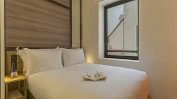 A neatly made bed with pillows, a bedside table with a lamp, and a window with a view of a building.